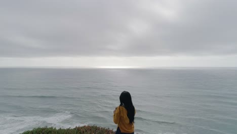 reveal of woman standing at coastal cliff on the pacific taking photos with phone reverse dolly