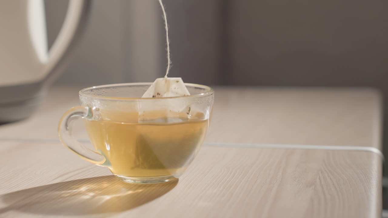 Hand reaching into a cup with tea bag floating inside, pouring hot water from kettle. Close-up of cup and tea bag steeping in hot liquid on kitchen table, preparing drink in calm morning setting