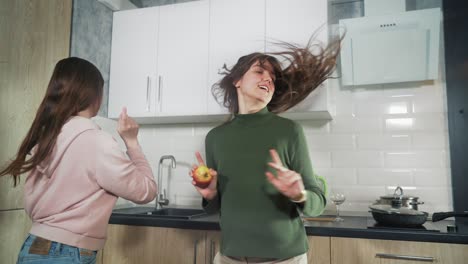 two happy young girls jumping and dancing in kitchen, cheerful friends or family having fun together at home