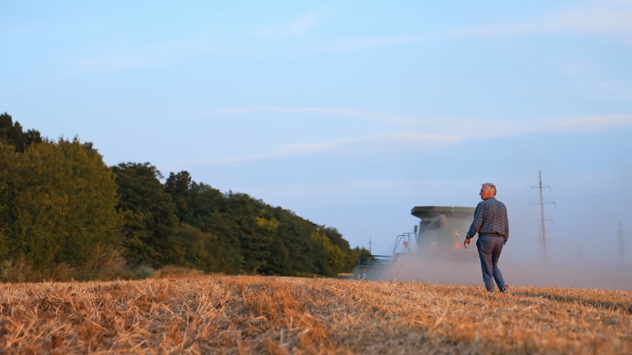 Slim man wearing a shirt and trousers follows the combine harvester working in the field. Season of bread harvesting