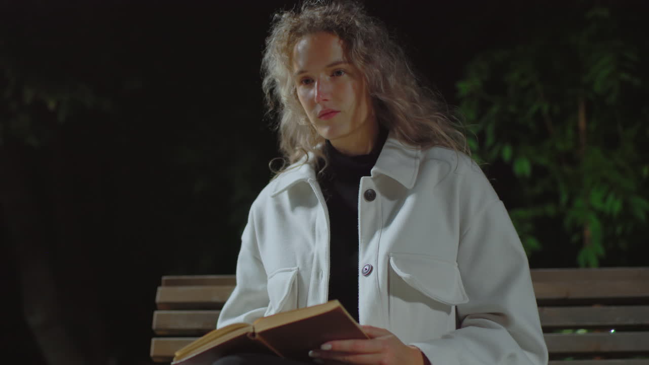 Thoughtful woman seated on bench at night reading book while gentle wind moves her curly hair under soft lighting, surrounded by dark greenery in peaceful and quiet outdoor park setting
