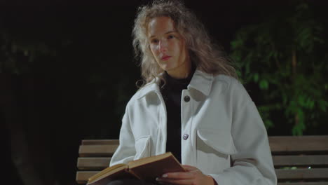 thoughtful woman seated on bench at night reading book while gentle wind moves her curly hair under soft lighting, surrounded by dark greenery in peaceful and quiet outdoor park setting