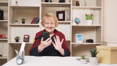 mujer de edad avanzada sonríe alegremente mientras habla por video llamada en su teléfono inteligente mientras está sentada en la mesa