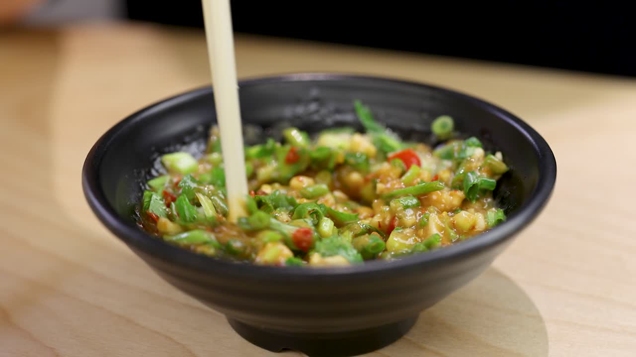 Chopsticks stir vibrant chili, garlic, and herbs in a black bowl under soft, even lighting