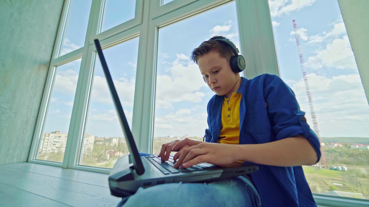 Teenager in headphones with a laptop by the window. Concentrated boy pressing buttons on a laptop while playing at home. Leisure time.
