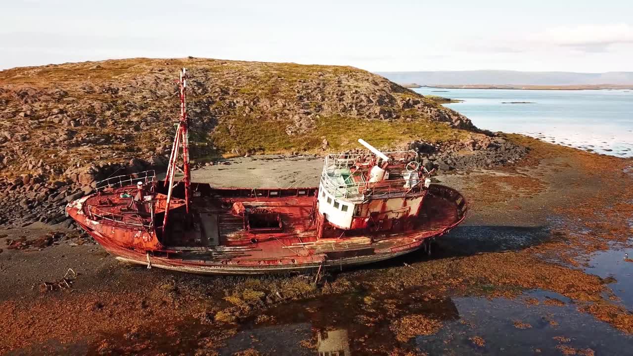 An aerial view of a rusted, abandoned shipwreck stranded on the rugged shores of Breidhabolsstadhur, Iceland, surrounded by rocky terrain and coastal waters under a clear sky