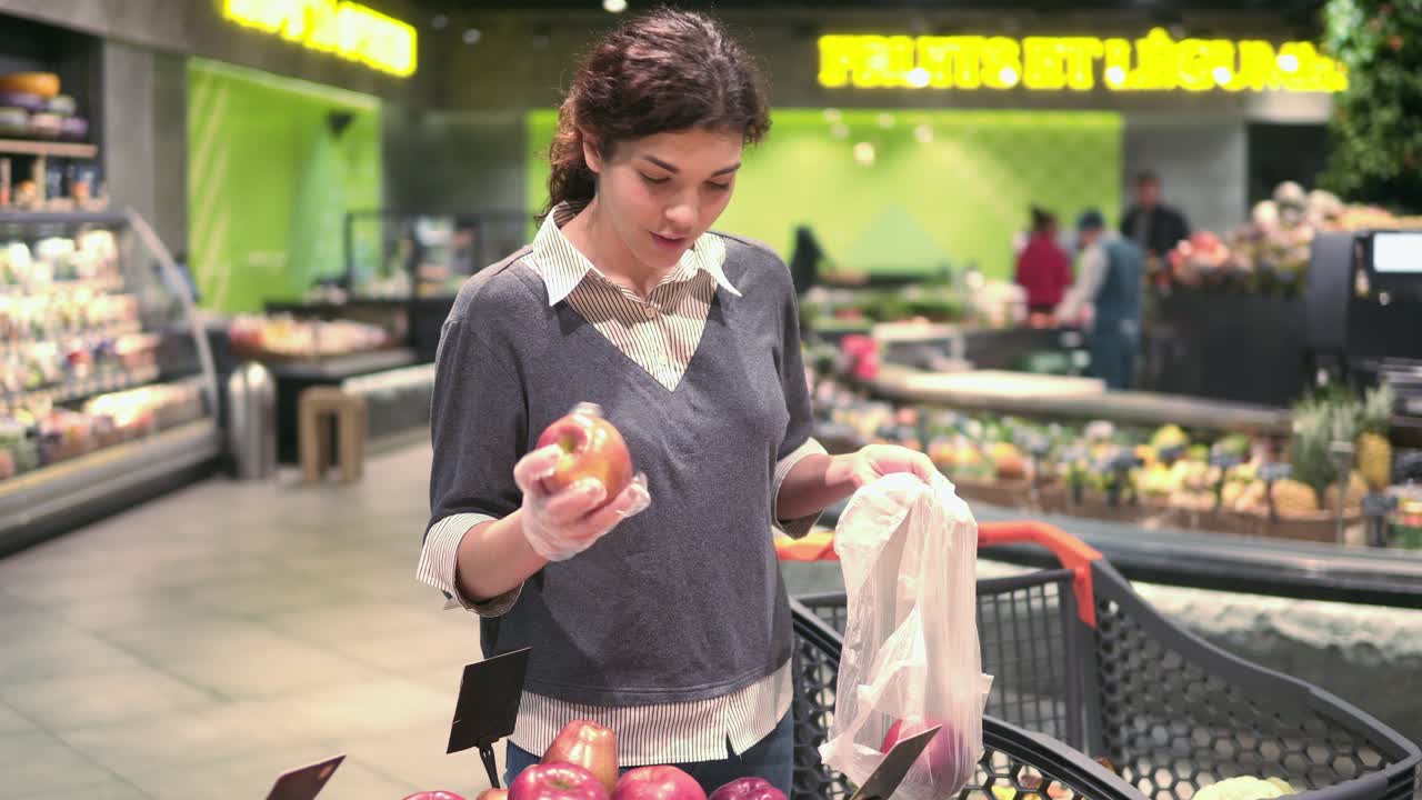 Young beautiful brunette girl in her 20's picking out red apples into a plastic bag at the fruit and vegetable aisle in a grocery store. Frontside footage.