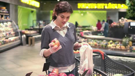 una joven hermosa chica morena de unos 20 años recogiendo manzanas rojas en una bolsa de plástico en el pasillo de frutas y verduras de una tienda de comestibles.