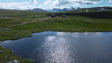 Aerial drone footage gliding over gorgeous green vibrant Sweden landscapes that has a small blue pond or lake that has water ripples on it. Large mountains with snow visible in the background. roads.
