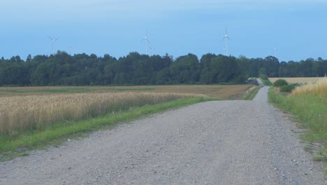 Granja-De-Turbinas-Eólicas-Que-Produce-Energía-Renovable-Para-Un-Mundo-Ecológico-Verde-En-La-Hermosa-Puesta-De-Sol,-Campo-De-Trigo-Dorado-Maduro-Y-Camino-De-Grava-En-Primer-Plano,-Plano-General