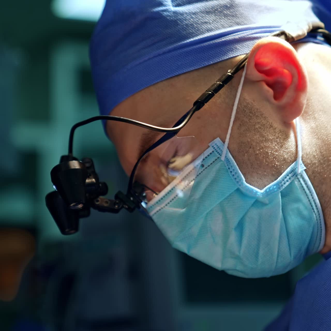 Male mid-aged surgeon in mask, cap and device glasses. Side view portrait of a doctor using metal instruments. Close up