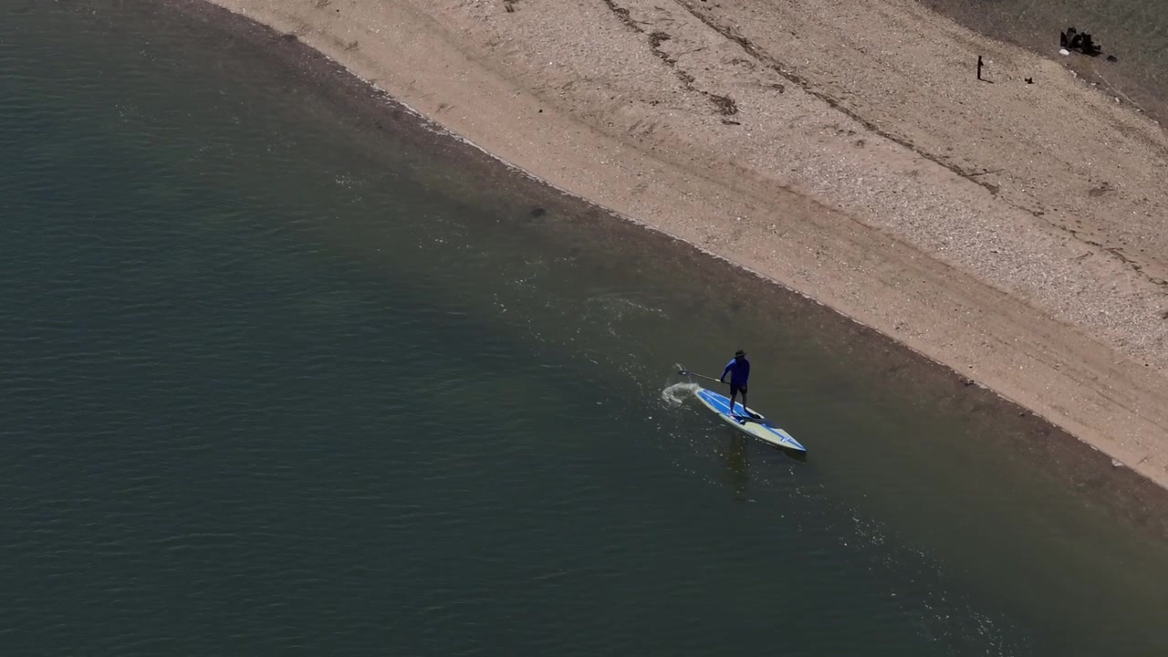 Una vista aerea di una persona che si gode una giornata di sole su una tavola da stand-up paddle in acque poco profonde vicino a un banco di sabbia. La telecamera del drone è inclinata verso il basso e si sposta a destra seguendo la persona dall'alto.