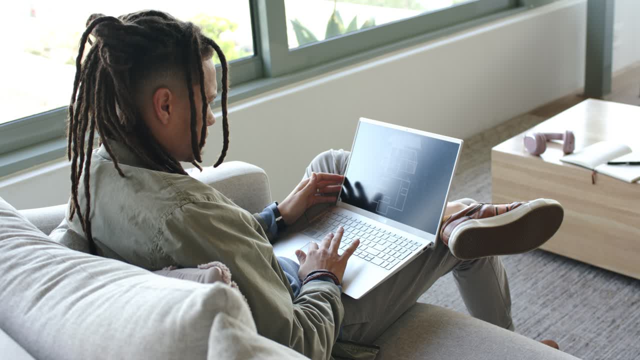 Individual opening silver laptop on couch by window and reviewing floor plan for design