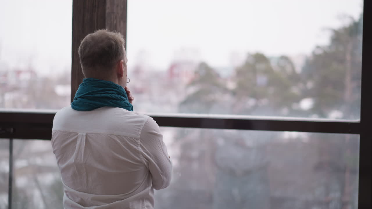 사람은 창문으로 겨울의 그림을 흡수합니다. 집중된 사람은 집의 평화로운 고립에서 문제를 해결하는 것에 대해 생각하고 있습니다. 평온한 고독.