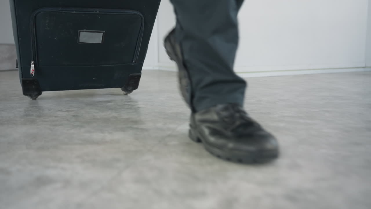 Low angle view captures traveler in motion pulling wheeled suitcase across smooth floor with black shoes in focus and blurred travel bag alongside open door in contemporary indoor setting