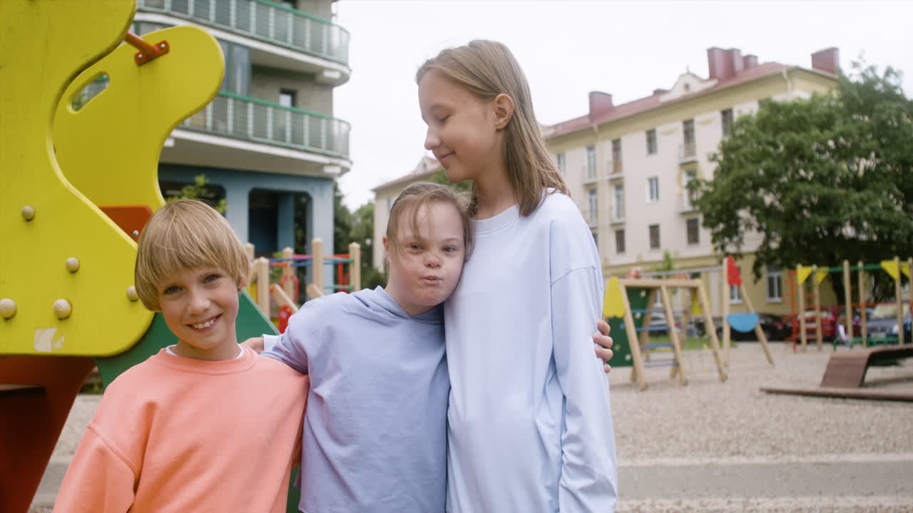 Close-up view of a little girl with down syndrome hugging another girl and little boy in the park on a windy day. They are looking at camera