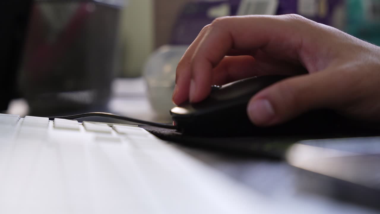 A cinematic close-up of a person's hand using a black computer mouse on a desk next to a white keyboard. Ideal for technology, business, and remote work themes