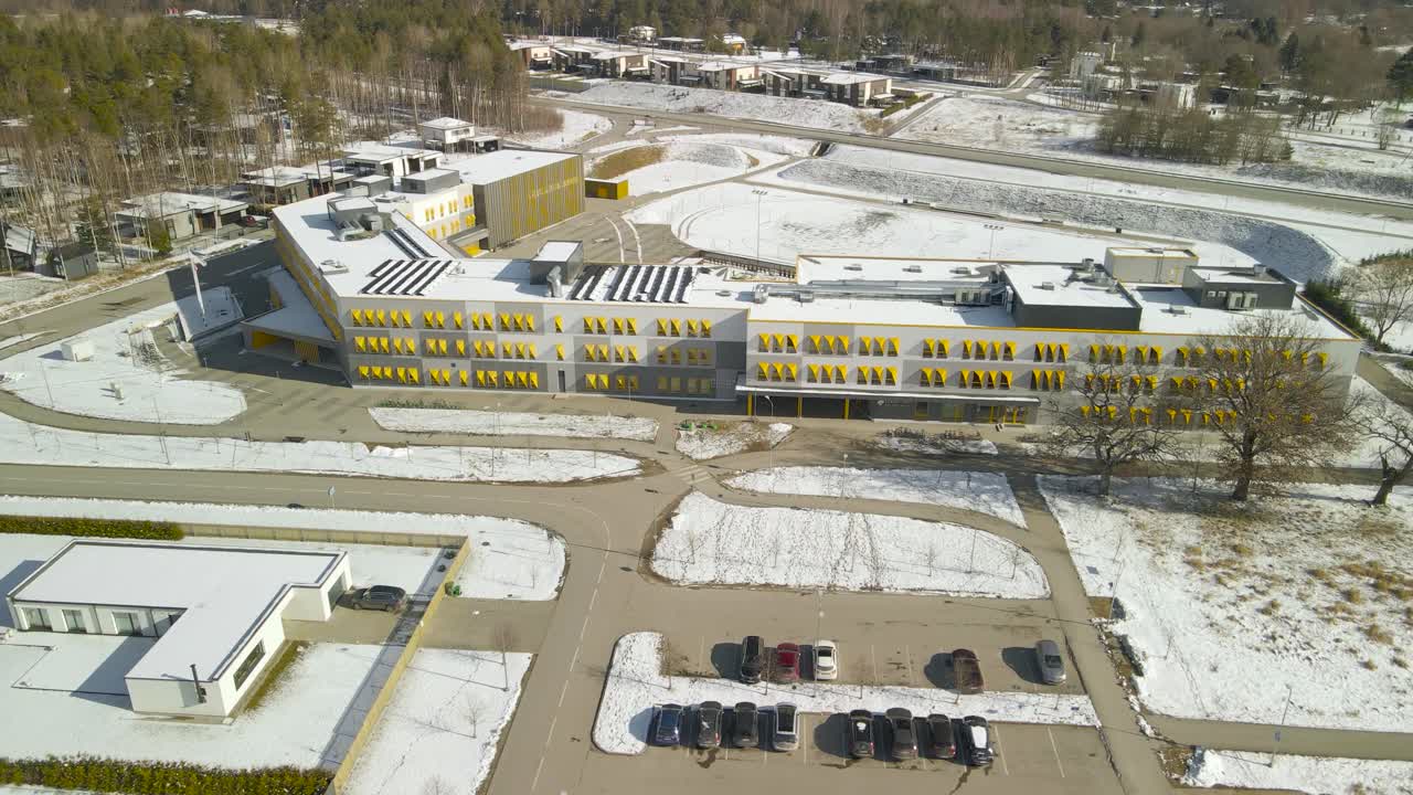 Aerial drone footage of a large modern and new architecture school called Möldre maja in Laagri Estonia during a sunny winter day while white snow is surrounding the school house. Cars parked outside.