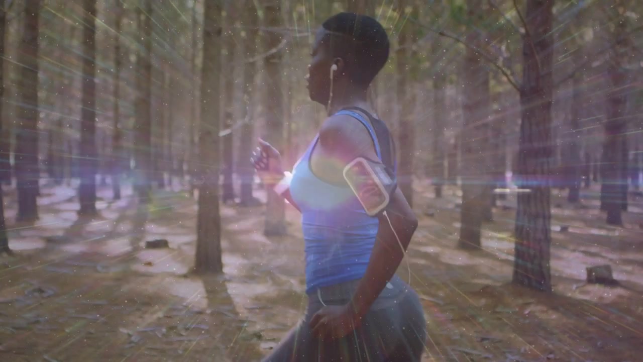 Corritrice che entra da destra, facendo jogging nella foresta, con la fascia da braccio per telefono che si sincronizza con un bagliore dell'obiettivo per il fitness