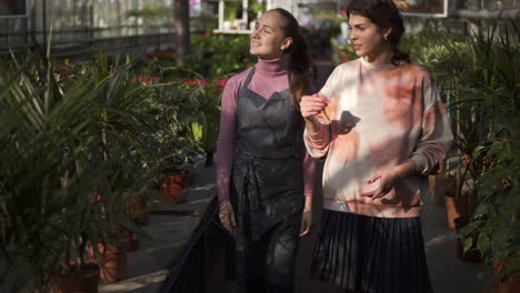 Young-smiling-female-florist-walking-with-her-boss-and-showing-her-different-plants,-explaining-information.-Young-woman-is-listening-carefully-to-the-florist