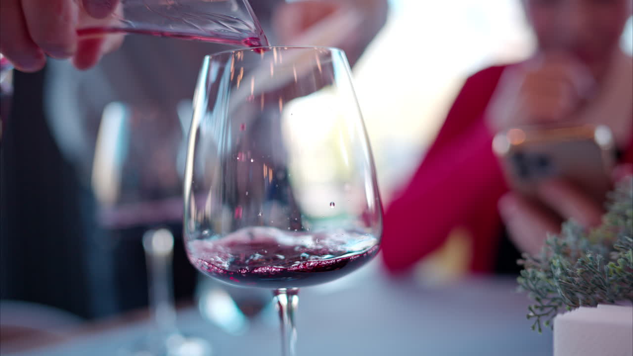 Red wine being poured in a glass for tasting at a restaurant table with blurred people on the background