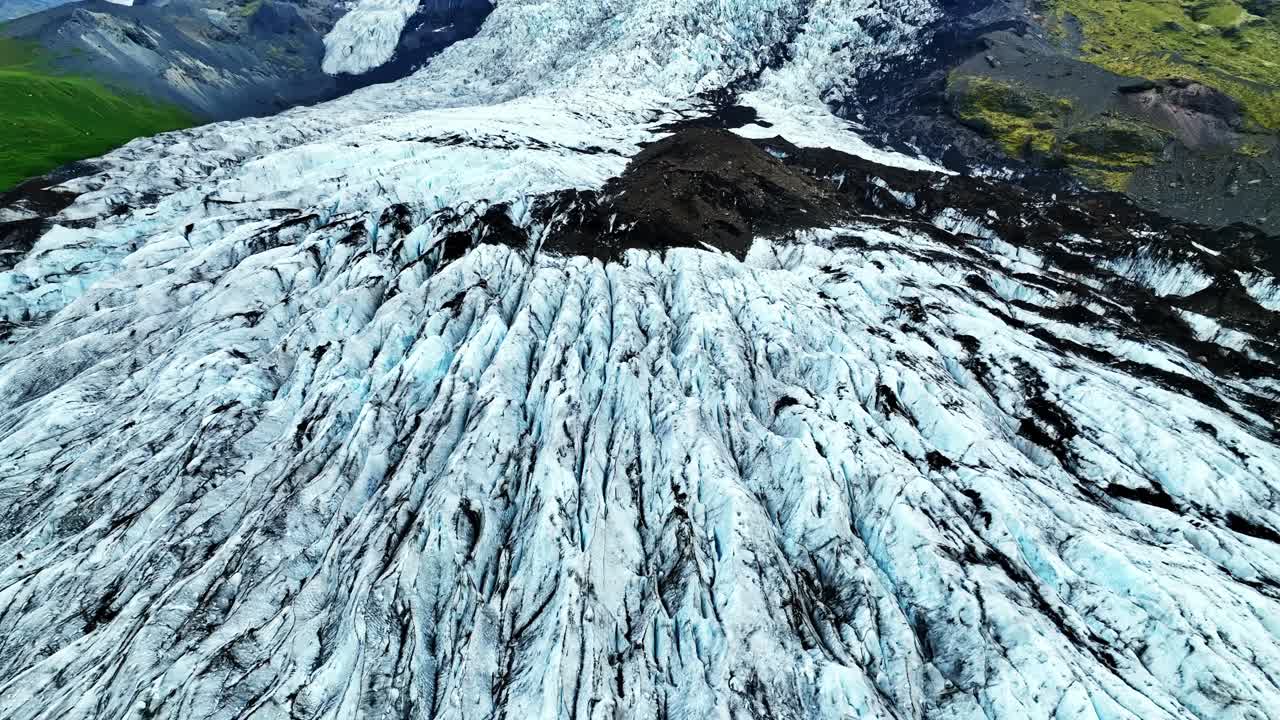 Dronebillede af en gletsjer, der flyder ned ad bakke med dybe isrygge, mørke vulkanske striber og et robust terræn, der danner en dramatisk naturlig komposition.