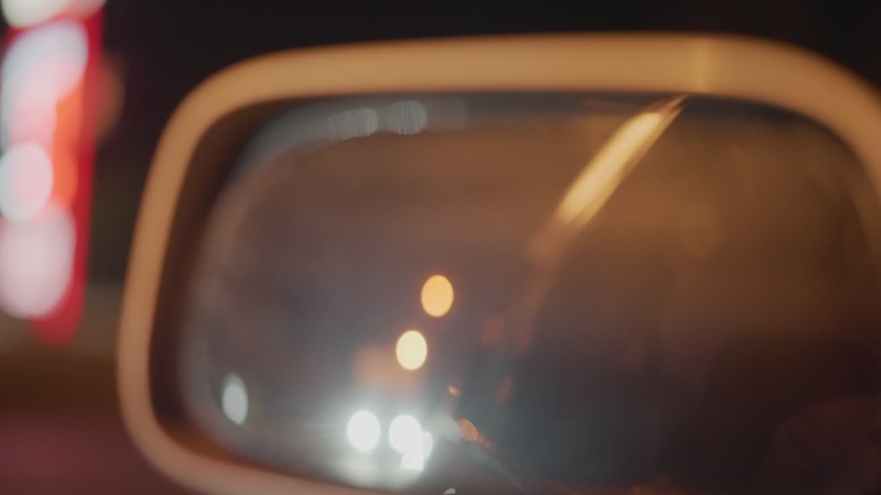 Espejo nocturno que refleja una luz distante, perspectiva del conductor en una carretera urbana, rastro de limpiaparabrisas difuminado en el cristal, faros borrosos y bokeh de lámparas, ambiente melancólico, indicios de tráfico y soledad silenciosa