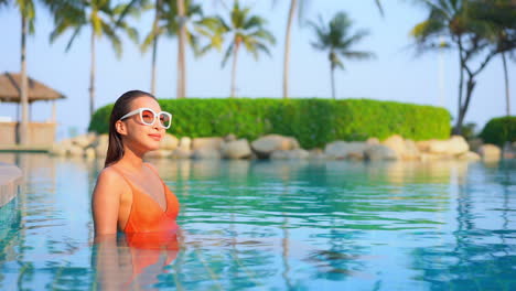 Asian-girl-in-bright-orange-swimsuit-turning-her-sight-while-inside-swimming-pool-of-exotic-tropical-resort-in-Hawaii