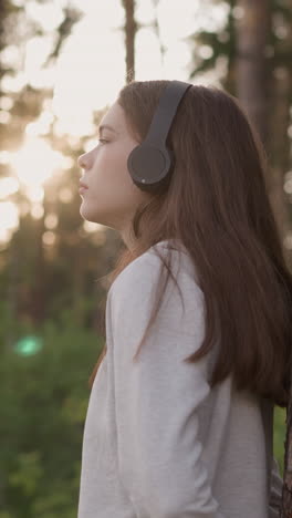 woman with headphones leans on tree trunk in forest. young lady listens to melancholy music thinking about bitter experience at sunset. feeling of solitude