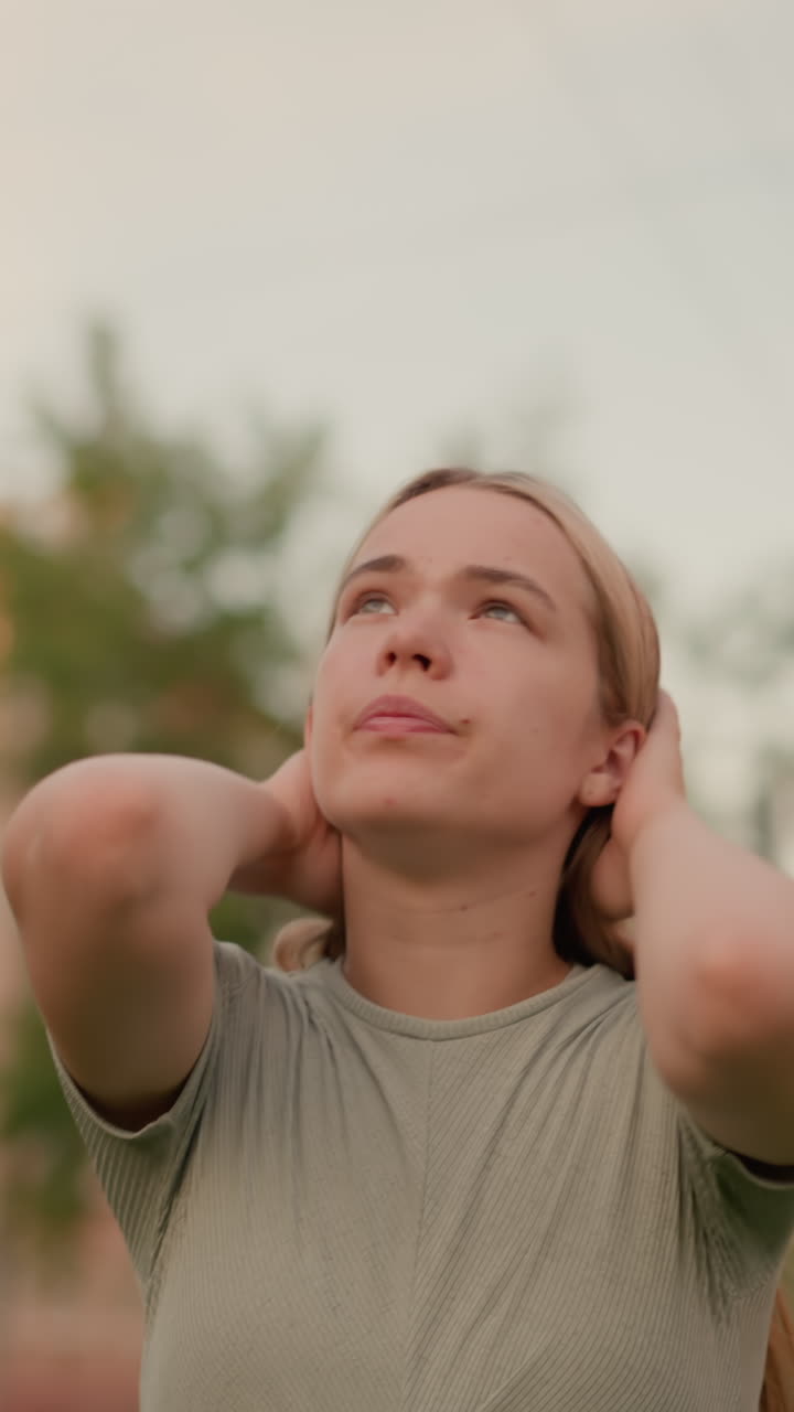joven al aire libre con las manos en la cabeza, mirando hacia arriba con una expresión contemplativa y de mal humor, rodeada de vegetación borrosa y estructuras urbanas