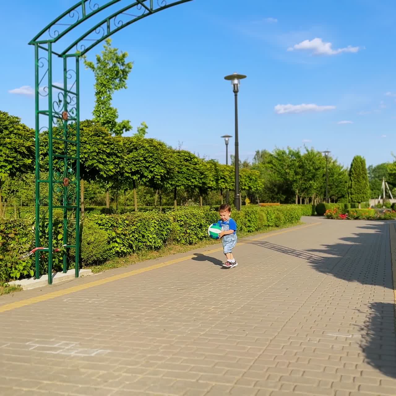 Lindo niño pequeño con un peto vaquero recoge una pelota y corre con ella. Un niño encantador divirtiéndose en el soleado parque verde.