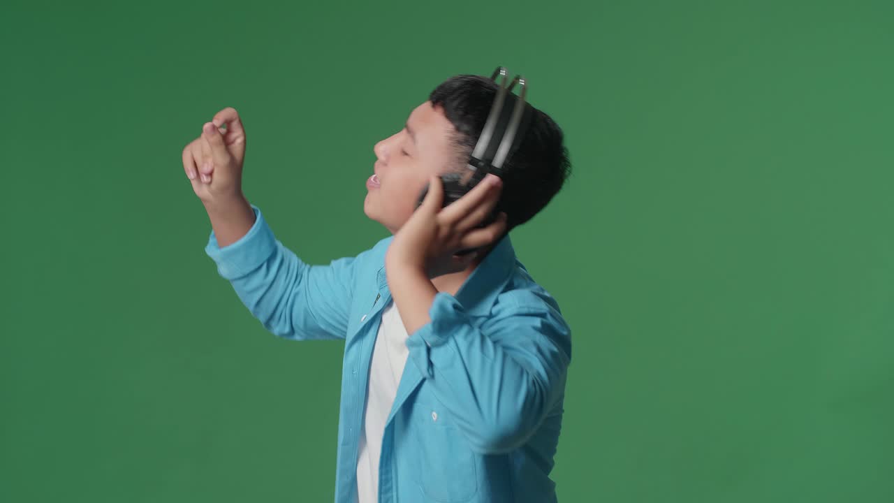 Side View Of Young Asian Boy Listening To Music With Headphones And Dancing In The Green Screen Studio