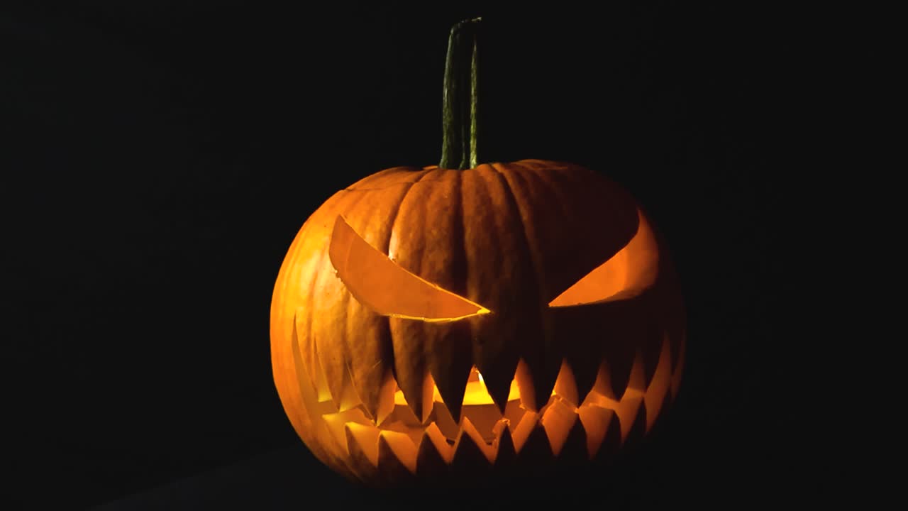 Video ravvicinato che orbita intorno a una zucca di Halloween arancione appena intagliata con un volto malvagio da Jack-o'-lantern. La zucca ha candele accese all'interno. Di fronte, sfondo nero da studio.