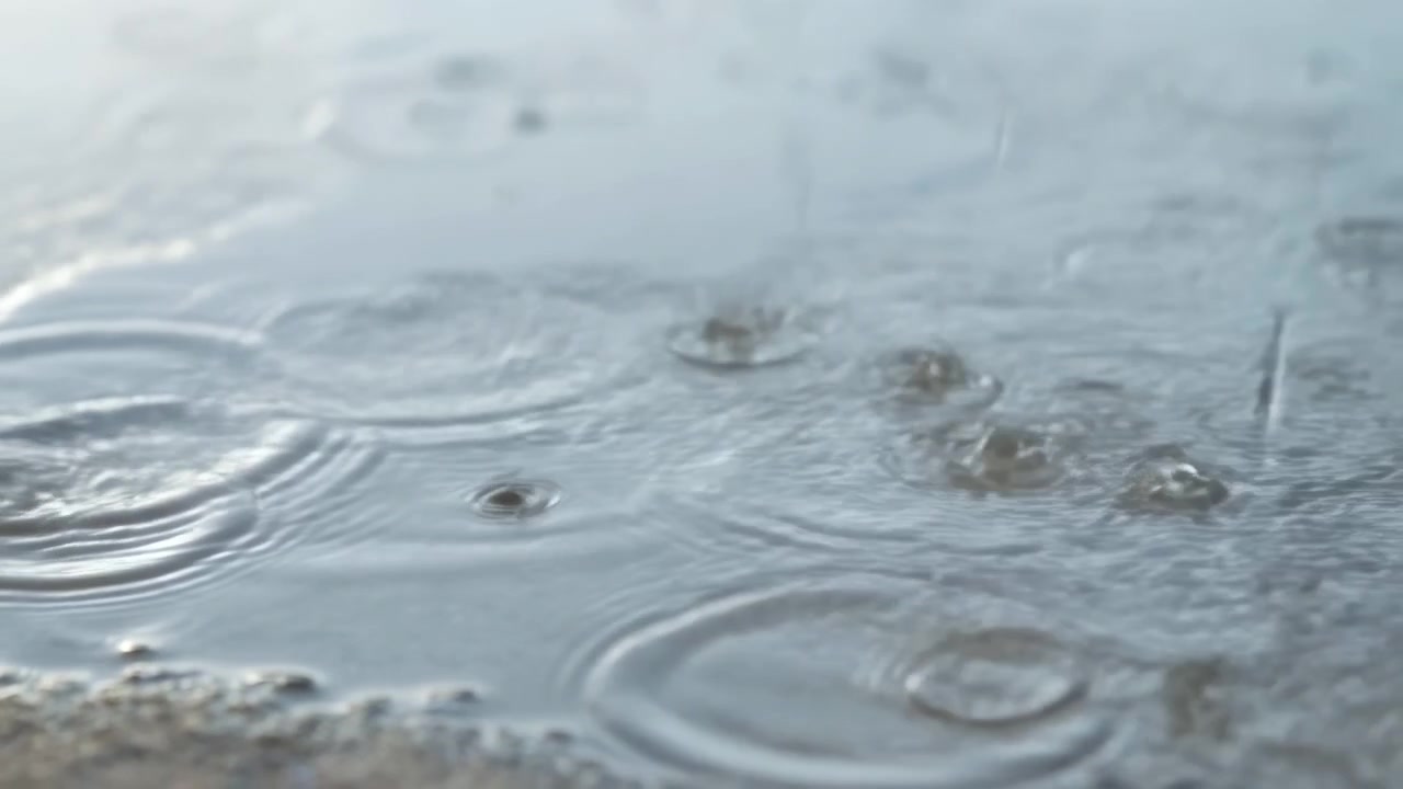 水たまりの表面に当たる雨粒が波紋を作る、柔らかな灰色の光、濡れた砂と同心円状の波紋のクローズアップ