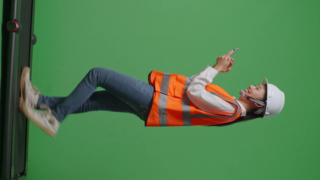 녹색 화면 배경 스튜디오에서 는 동안 스마트폰을 사용하여 안전 헬을 착용한 아시아 여성 엔지니어의 전체 몸의 측면 시각