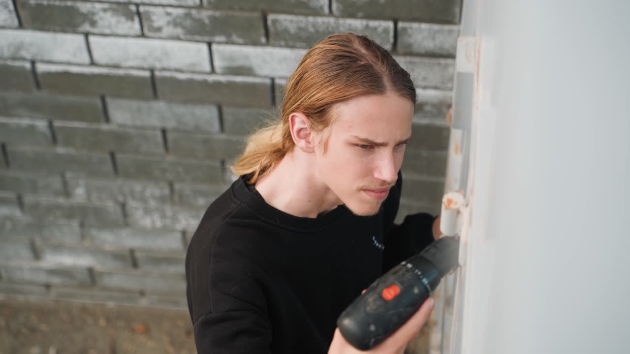 Young caucasian handyman drilling door hinge, focused expression near brick wall, using cordless screwdriver to install metal hinge on exterior door, wearing black sweater and tied back blonde hair