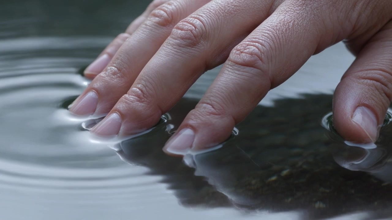 Touching adult hand pressing fingertips into calm pond, creating ripples near submerged rock