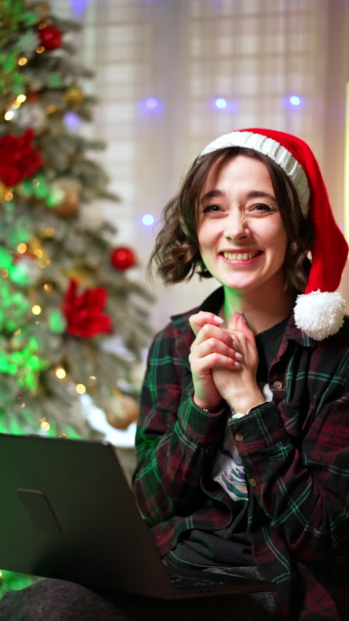 Happy rejoicing Caucasian girl in Santa cap. Cheerful lady is clapping hands looking at laptop. Christmas tree at backdrop. Vertical video.