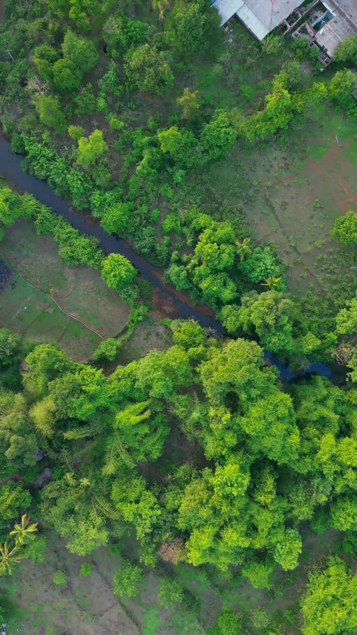 A drone view of Devrukh, Konkan, revealing a picturesque village among lush greenery and traditional houses. A winding river cuts through the serene landscape, showcasing rural charm.