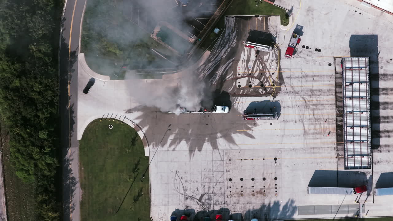 Drone view of a semi-truck fire near a parking lot as smoke billows upward while emergency vehicles respond to the scene