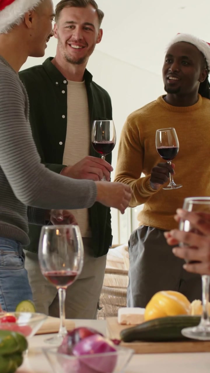 Vertical video: Taking offered wine, man in green shirt toasting at kitchen island with Santa hats