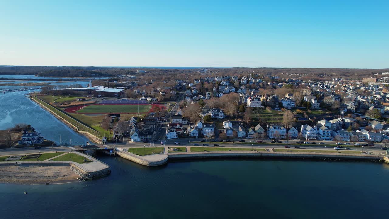 Vista de dron de la ciudad de Gloucester, Massachusetts, con frente marítimo soleado, agua azul del puerto, barrios y costa pintoresca, creando una atmósfera clásica de ciudad costera de Nueva Inglaterra en un día despejado. Panorámica amplia lateral.