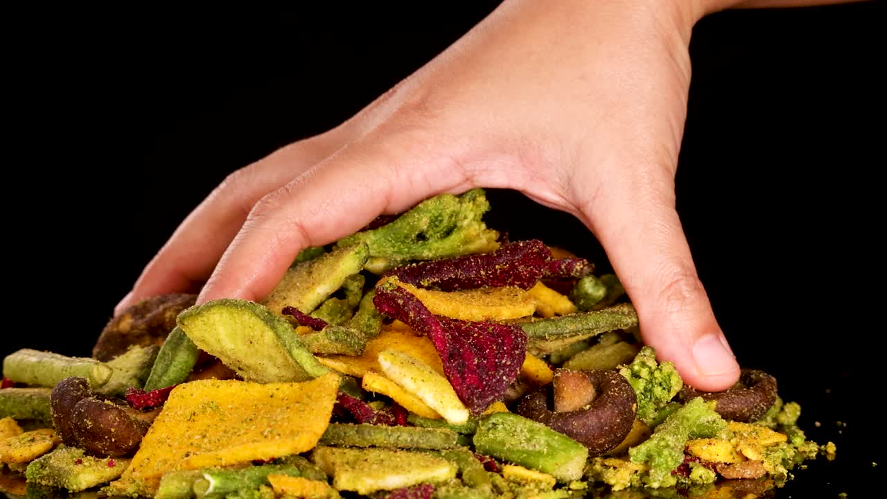 Hand selects assorted dried vegetable chips, crisp texture, vibrant colors, studio lighting, black backdrop