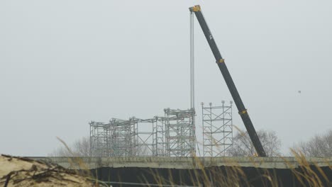 Intensive-work-on-the-construction-site-located-near-the-coast-of-the-sea,-mobile-crane-moving,-overcast-day-with-fog,-distant-medium-shot