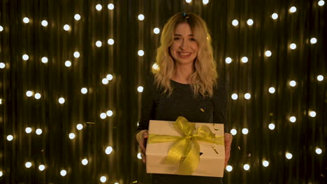 young beautiful winner female holding a gift box with golden bow, shaking hair while falling glitter confetti