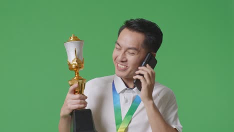 금메달을 가진 아시아인 남자가 금 트로피를 들고 스튜디오에서 녹색 화면 배경에서 스마트폰으로 이야기하고 있습니다.