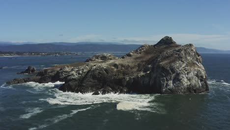 Powerful-aerial-drone-footage,-slowly-approaching-a-very-large-Pacific-ocean-boulder-which-is-surrounded-by-crashing-waves