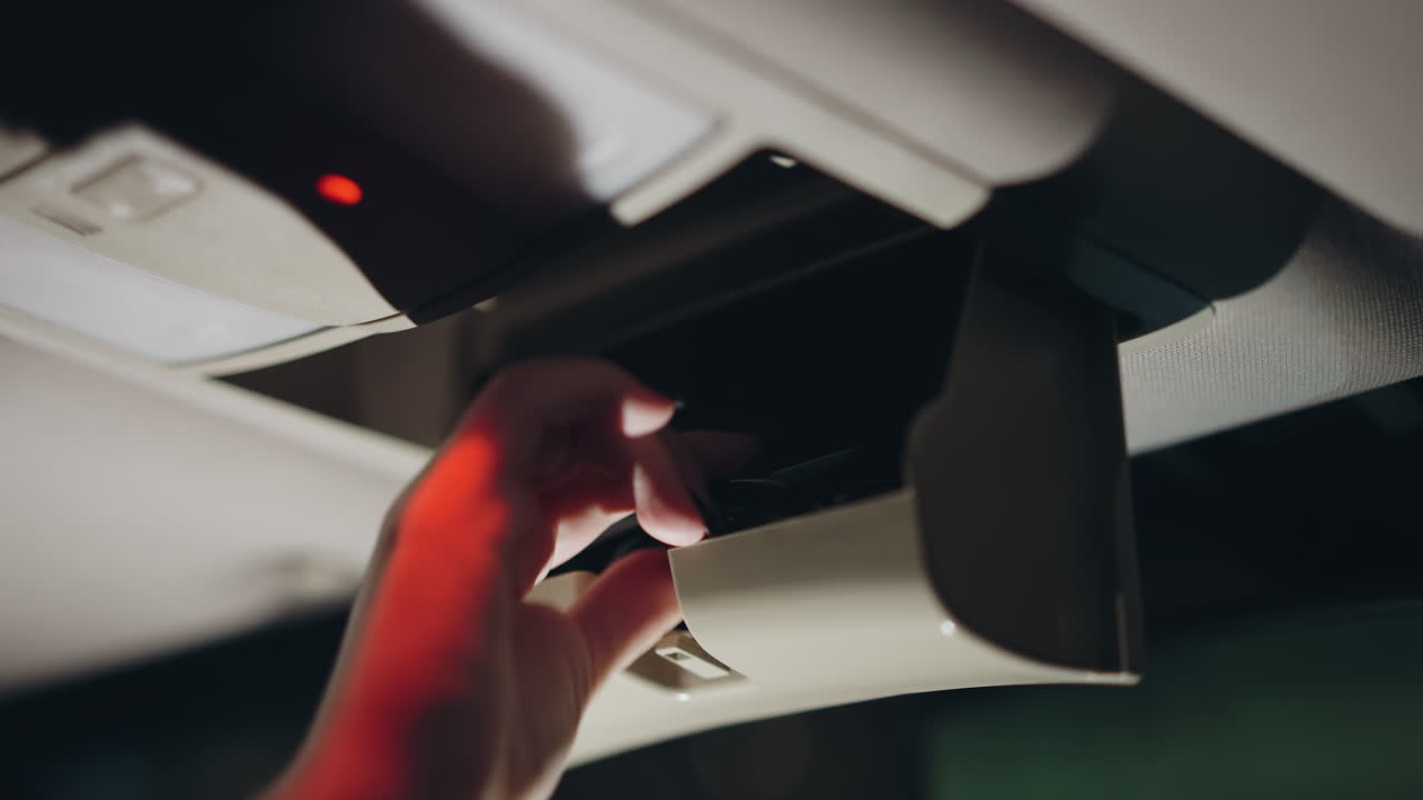 vista en primera persona de una mujer abriendo una caja del techo del coche y colocando gafas en el interior por la noche. la escena destaca su mano con uñas pintadas de negro mientras interactúa con la caja del tejado del coche