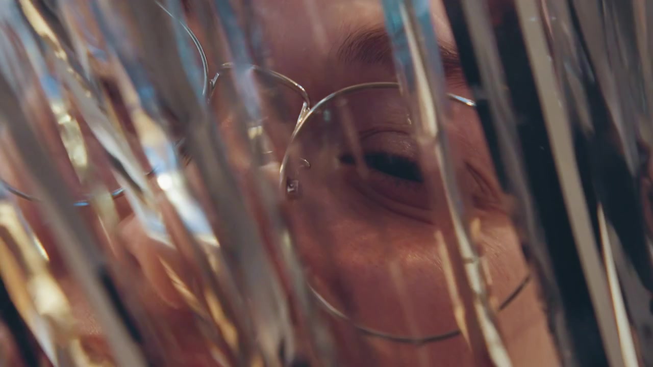 Closeup face behind metallic tinsel curtain, glasses and eye partially visible through shimmering foil strands, soft bokeh and reflective highlights create intimate mysterious mood, studio lighting