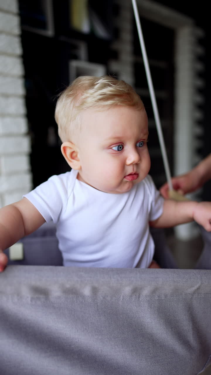 Beautiful little baby with blond hair and plump cheeks sitting in a swing. Active time of a kid at home. Vertical video.